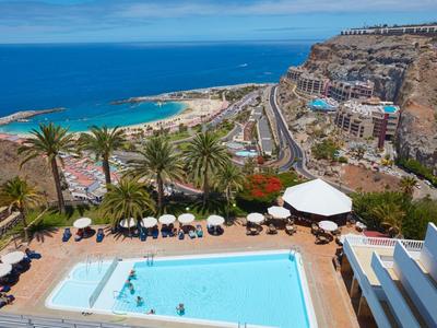 Blick auf Pool mit Sonnenschirmen, Palmen und Küste mit Strand unter blauem Himmel.