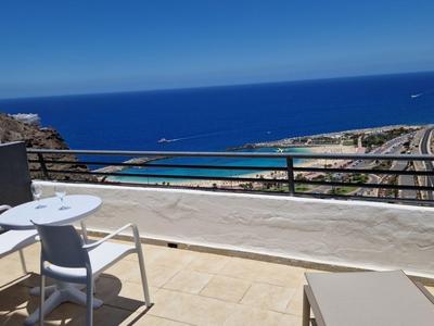 Balkon mit weißen Stühlen und Tisch, Blick auf Meer und Küste bei klarem Himmel