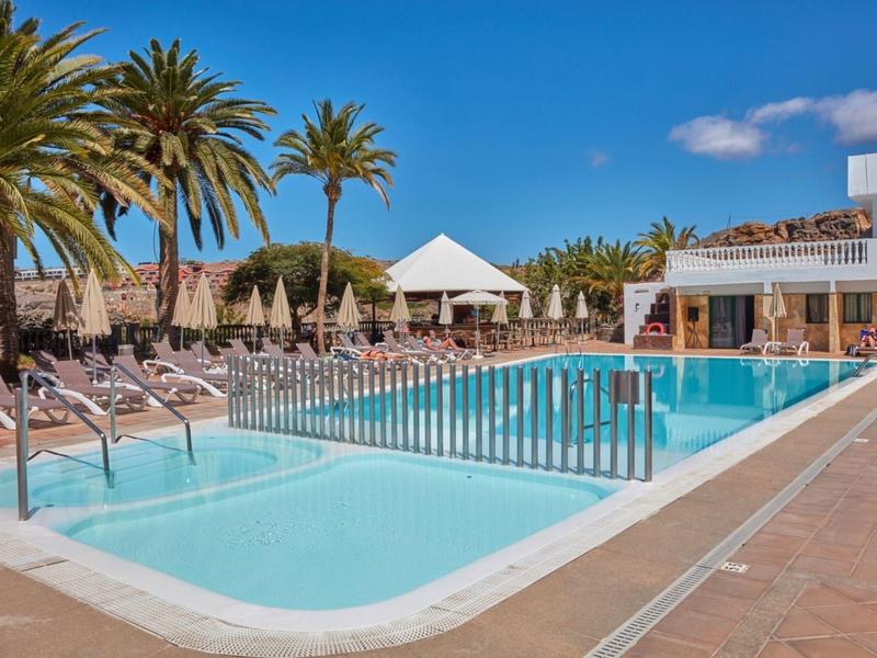 Großer Außenpool mit klarem Wasser, Liegestühlen und Palmen unter blauem Himmel.