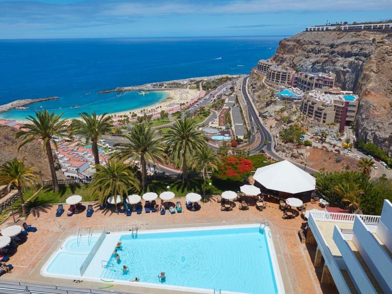 Blick auf Pool mit Sonnenschirmen, Palmen und Küste mit Strand unter blauem Himmel.