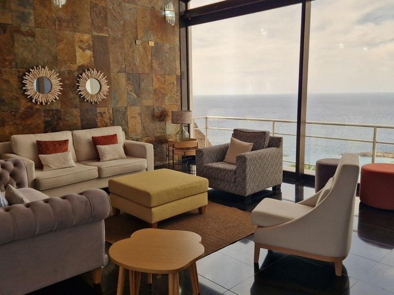 Modern eingerichtetes Wohnzimmer mit Sofa, Sesseln, Hocker und großem Fenster mit Meerblick.