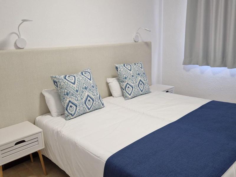 Helles Schlafzimmer mit großem Bett, blauem Tagesdecke, gemusterten Kissen und weißem Nachttisch.