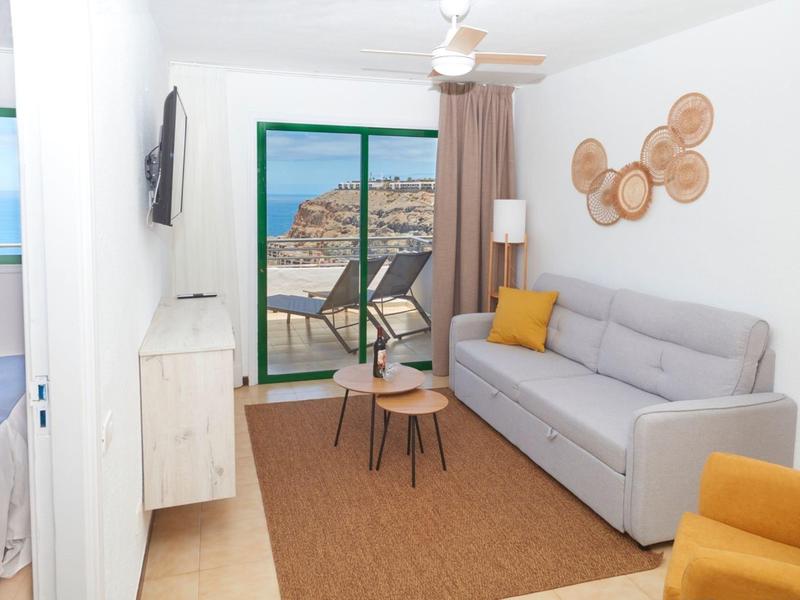 Helles Wohnzimmer mit grauem Sofa, rundem Teppich, Balkon und Aussicht auf Meer und Felsen.