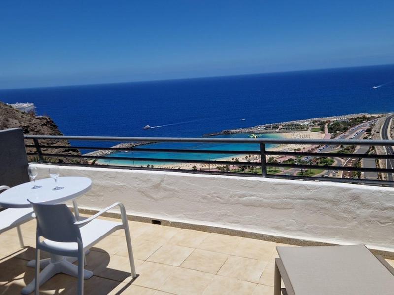Balkon mit weißen Stühlen und Tisch, Fliesenboden, Metallgeländer, Blick auf Meer und Hügel.