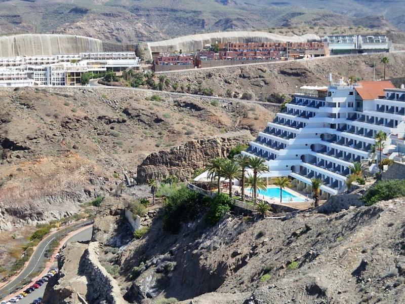 Häuser und Pools auf felsigem, trockenem Gelände mit Bergen im Hintergrund bei sonnigem Wetter.