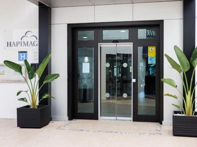 Eingang eines Hotels mit automatischen Glasschiebetüren und zwei großen Pflanzen in schwarzen Töpfen.