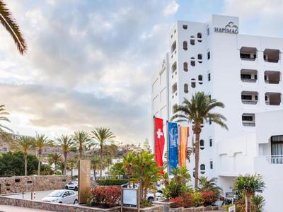 Weißes Hotelgebäude mit Palmen und bunten Flaggen bei bewölktem Himmel