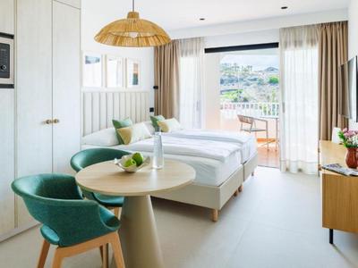 Helles Hotelzimmer mit Doppelbett, grünem Sessel, Holztisch und Balkon mit Aussicht.