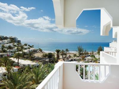 Blick von einem Balkon mit weißem Geländer auf Palmen, Gebäude und das Meer bei sonnigem Wetter.