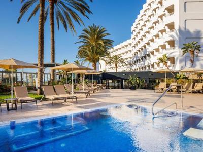 Hotelpool mit Liegestühlen, Sonnenschirmen und Palmen unter klarem Himmel.