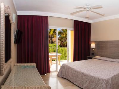 Chambre avec lit double, ventilateur de plafond et vue sur terrasse avec jardin.