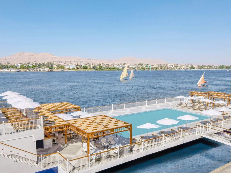 Piscina che si affaccia su un lago con ombrelloni e vele sullo sfondo.