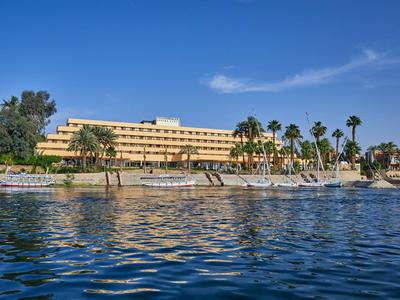 Großes gelbes Hotel am Ufer mit Palmen, blauem Himmel und ruhigem Wasser im Vordergrund.