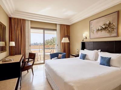 Elegantní hotelový pokoj s velkou postelí, křeslem, psacím stolem a balkonem s výhledem na pláž.