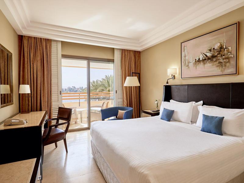 Elegantní hotelový pokoj s velkou postelí, křeslem, psacím stolem a balkonem s výhledem na pláž.