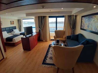 Helles Hotelzimmer mit Sofa, Sesseln, Teppich, TV, Bett und Balkon mit Meeresblick.