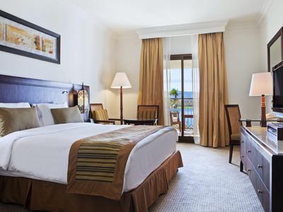 Helles Hotelzimmer mit großem Bett, Schreibtisch, Fernseher und Balkon mit Blick auf Wasser.