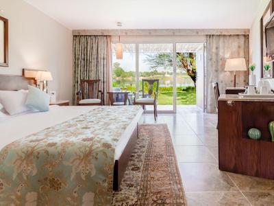 Helles Hotelzimmer mit Doppelbett, Teppich, Schreibtisch und Blick auf Garten durch große Fenster.