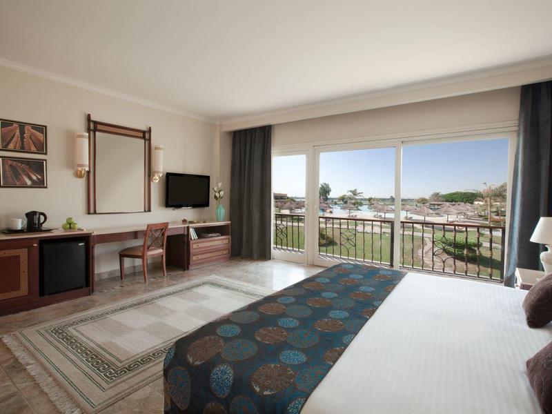 Bright hotel room with large bed, rug, desk, and balcony overlooking the city.