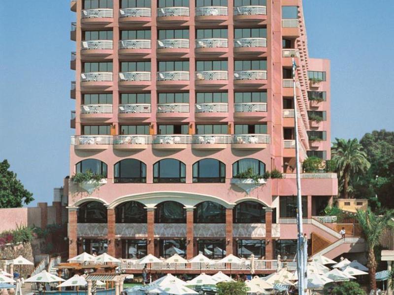 Edificio de hotel rosa de varios pisos con balcones y piscina al aire libre junto al agua.