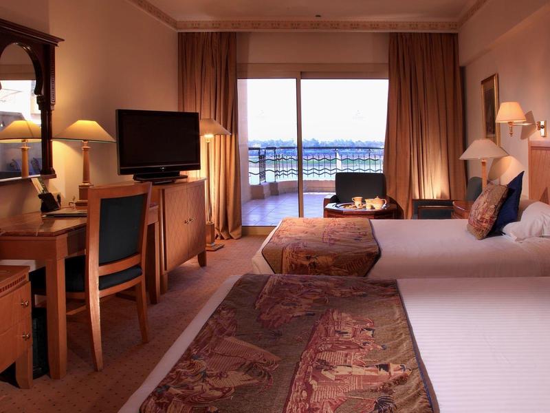 Camera d'hotel con due letti, scrivania, TV e balcone con vista sull'acqua.