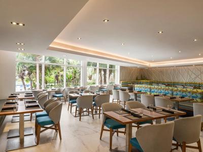 Moderner, heller Speiseraum mit großen Fenstern, Holz- und Polstermöbeln, gedeckten Tischen, Pflanzendekor.