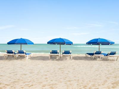 Drei blaue Sonnenschirme und Liegen am Sandstrand vor ruhigem Meer und blauem Himmel.
