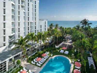 Hotel mit großem Pool, umgeben von Liegestühlen und Palmen, Meer im Hintergrund unter klarem Himmel.