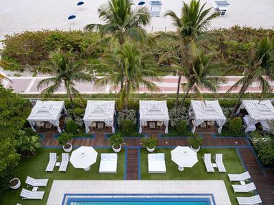 Blick auf einen rechteckigen Pool mit Liegestühlen, Palmen, Strand und Meer im Hintergrund.