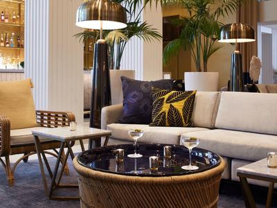 Gemütliches Wohnzimmer mit beigem Sofa, braunem Sessel, rundem Tisch und großen Pflanzen im Hintergrund.