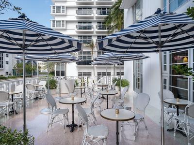 Terrassenbereich mit weißen Metallstühlen und Tischen, blau-weiße Sonnenschirme, moderner Gebäudefassade.