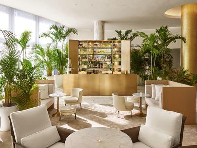 Moderner Gastraum mit weißen Sesseln, runden Tischen, goldfarbenem Tresen und vielen grünen Pflanzen.