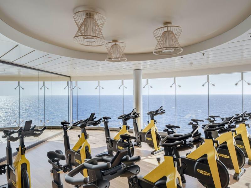 Ein moderner Fitnessraum mit gelben Spinning-Fahrrädern vor großen Panoramafenstern mit Blick auf das Meer.