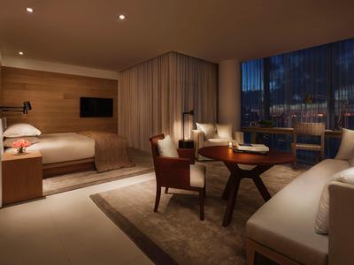 Modernes Hotelzimmer mit Wohnbereich, Bett, Fernseher und großer Fensterfront bei Abenddämmerung.