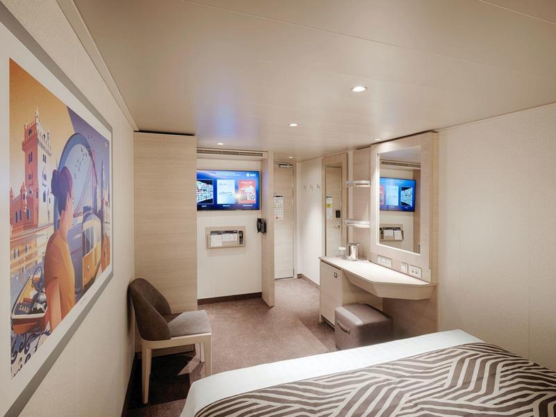 Kompaktes Hotelzimmer mit Bett, Stuhl, Schreibtisch, Spiegel und einem Fernseher an der Wand.