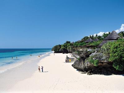 Breiter Sandstrand mit klar blauem Wasser, Felsen und grüner Vegetation bei Sonnenschein