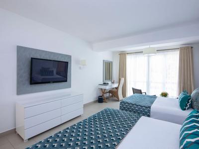 Helles Hotelzimmer mit zwei Betten, TV und Schreibtisch an einer Fensterfront.