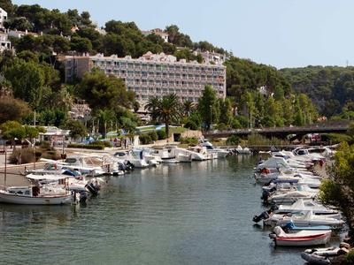 Harbor with docks and yachts in front of a hotel building on a wooded hillside.