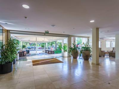 Bright hotel lobby with large plants, glass walls, and seating areas overlooking terrace.