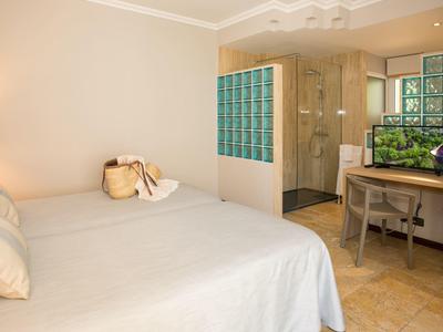 Helles Hotelzimmer mit Doppelbett, Schreibtisch und Blick auf eine Duschkabine mit Glasbausteinen.