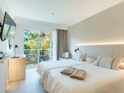 Helles Hotelzimmer mit zwei Betten, Fernseher und Balkon mit Blick auf Grünflächen.