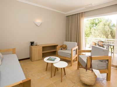 Helles Wohnzimmer mit Sofa, Sesseln, Couchtischen und Balkon mit Blick auf grüne Bäume.