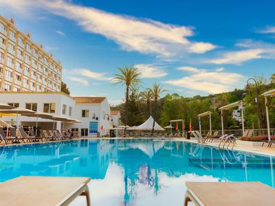 Großer Pool mit Liegestühlen vor einem Hotel unter blauem Himmel.