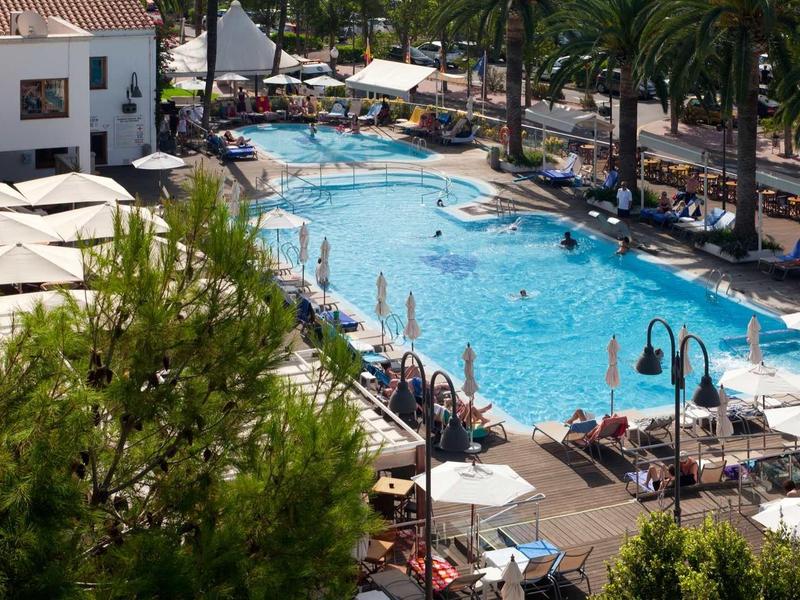 Large hotel pool with sun loungers, umbrellas, and surrounding palm trees on a sunny day.