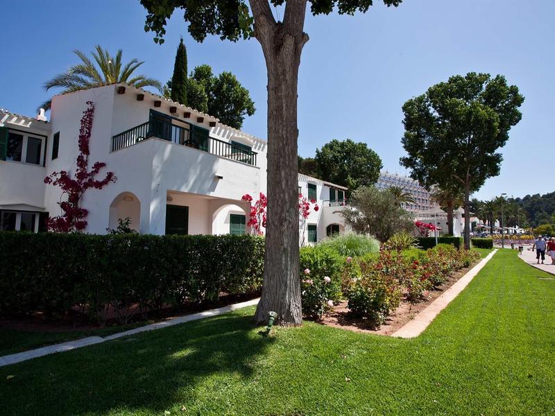 White holiday houses with balconies beside a well-kept lawn and trees under a blue sky.