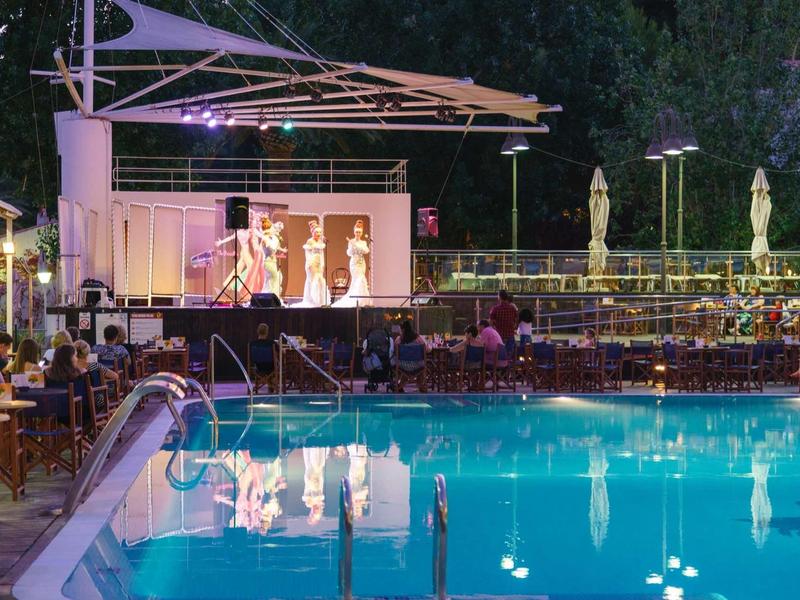 Illuminated stage show with dancing performers beside a pool at a hotel at night.