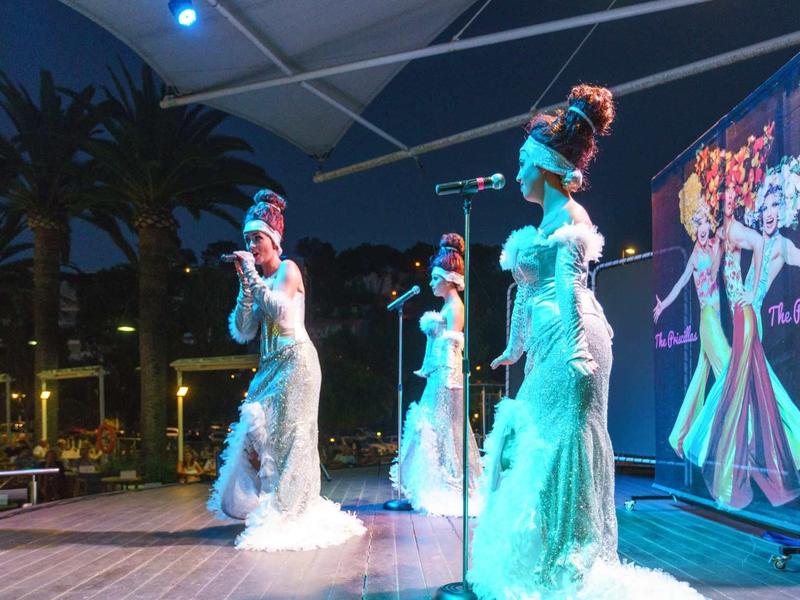 Three women in white dresses perform on a stage at night with palm trees in the background.
