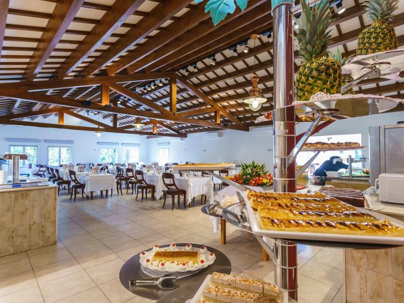 Bright restaurant with wooden beam ceiling, tables, chairs, and buffet with pastry and pineapples.
