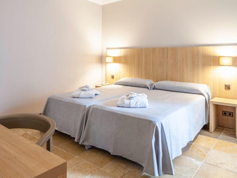 Helles Hotelzimmer mit zwei Einzelbetten, grauen Bettdecken, Nachttischlampen und einem Sessel.