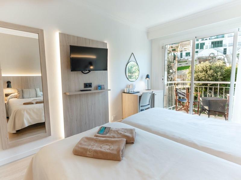 Helles Hotelzimmer mit Doppelbett, Wandspiegel, TV und Balkon mit Stadtblick.